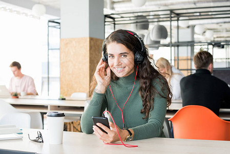 当代年轻女穿着大耳机复制空间在工作场所通过智能手机收听音乐时微笑着镜头的肖像背景图片