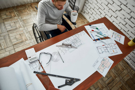 坐在轮椅上的年轻男建筑师坐在现代办公室的工作场所看蓝图和喝咖啡工程具残疾概图片
