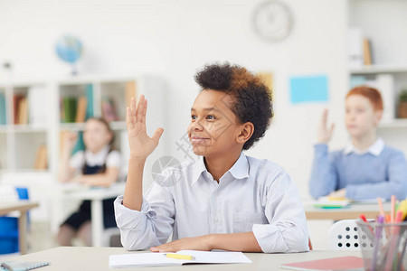 非洲微笑的男生坐在课桌前举手在学校上图片