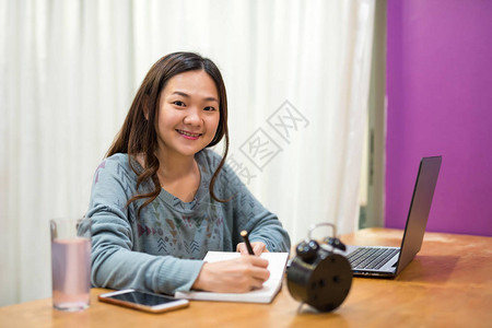 大学亚洲女学生的肖像在电脑笔记本电脑附近的工作台上写或记笔期末考试的快乐亚洲女孩讲背景图片