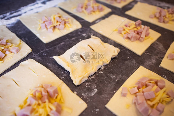 配有火腿和奶酪的酥皮点心放在一张撒有面粉的深色厨房桌子上包子在烹调过程中生菜家庭烘焙图片