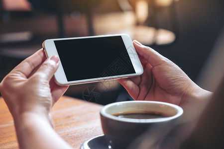 手握和使用带有空白黑屏的白色手机水平观看木桌上的咖啡杯的模型图像背景图片