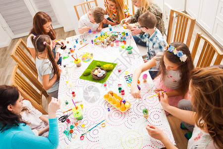 儿童坐在大桌旁装饰和绘画传统复活节鸡蛋的首版照片成年妇女帮助儿童创意讲习图片