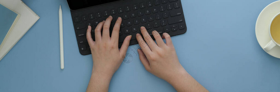 用茶杯和日记笔本在轻蓝桌的数码平板电脑上打字的女自由职图片