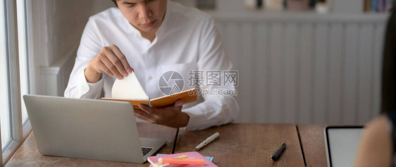 用笔记本电脑和笔记本在图书馆木制书桌上准备考试的年轻男大学生被剪图片