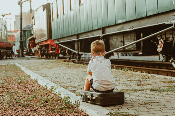 带孩子度假一个男孩坐在火车站月台上的手提箱上一列火车在等图片