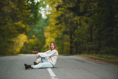 穿着牛仔裤和白毛衣的快乐女人坐在秋森林沥青路图片