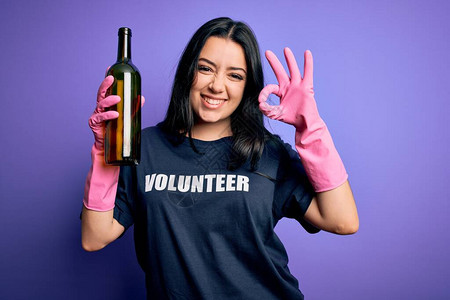 身穿志愿者T恤的褐发年轻女子在紫色背景上采摘玻璃瓶图片