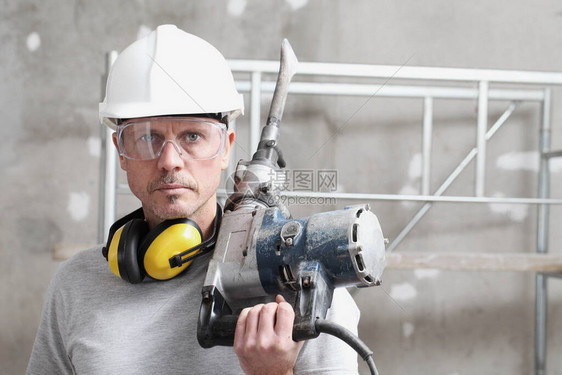 男子建筑工人的肖像与手提钻与安全帽听力保护耳机和防护眼镜看在内部建筑工地背景图片
