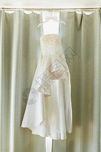 在窗帘背景下挂着新郎领结的衣架上装饰原设计新娘的白色礼服图片