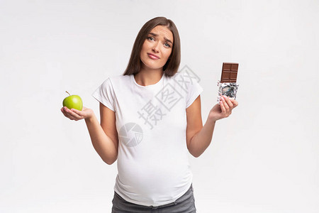 怀着苹果和巧克力的迷惑孕妇选择健康或不健康饮食在白背景的图片