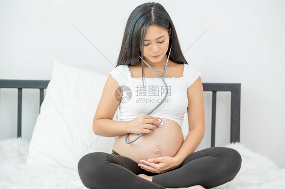 亚洲怀孕母亲的肖像在晨光下坐在床上时用听诊器听婴儿在子宫里的声音在健康母亲的子宫里照顾好婴儿的图片
