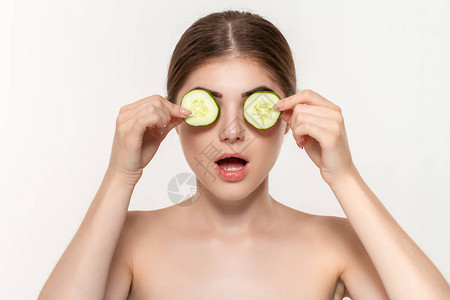 一个美丽的年轻女孩拿着绿色黄瓜片靠近眼睛孤立在白色背景的特写肖像美容图片