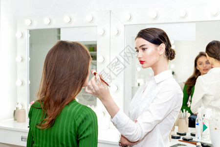 专业化妆师在美容院为年轻美女施用脸红的粉红色粉末图片