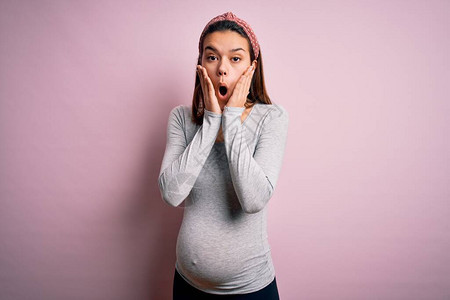 年轻漂亮的少女怀孕期待婴儿在孤立的粉红色背景中害怕震惊讶和惊讶的表情图片