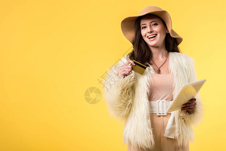 穿着毛皮外套的快乐优雅女孩戴着数字平板电脑和信用卡的帽子在图片