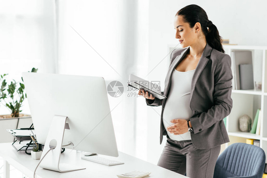 怀孕妇女用站在离桌近的办公室里时读笔图片