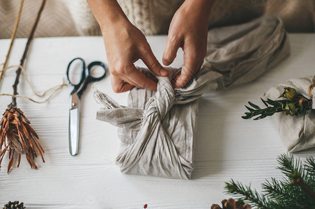 用天然冷杉草本植物松果和剪刀在白色质朴的桌子上用亚麻织物包裹圣诞礼物图片