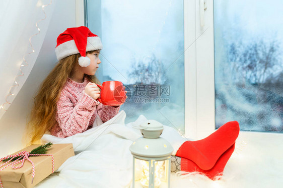 穿粉红色毛衣的可爱小女孩和圣塔帽坐在窗台上手上拿着一杯图片
