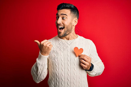 英俊的年轻帅哥将红形心脏作为浪漫和健康象征指向并用拇指举到一旁微笑图片