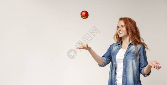 快乐的美丽红发女孩在光工作室背景上向空气中扔苹果图片