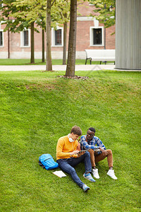 坐在校园绿草坪上并使用数字平板电脑复制空间的两名学生图片