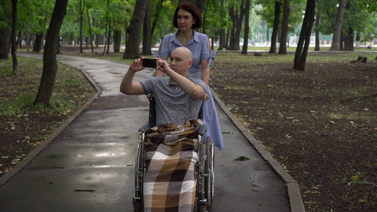 一名妇女带着一名坐在轮椅上的肿瘤科年轻男子穿过公园背景图片