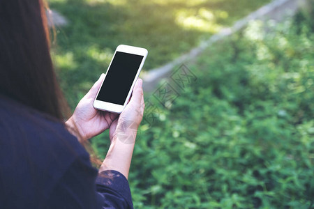 在绿色自然背景的户外手持和使用带有空白黑色桌面屏幕的白色智能手机的图片