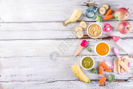 婴幼儿食品有蔬菜水果泥绿色橙色黄色的碗西兰花胡萝卜香蕉苹果带婴儿配饰和玩具木制白色背图片