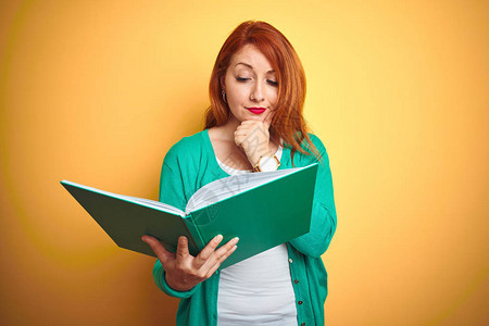 年轻的红发女学生在黄色孤立的背景下阅读绿皮书图片