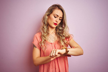 年轻漂亮的女人穿着T恤站在粉红色的孤立背景上查看手表上的时间图片