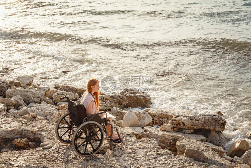 一位年轻的红发残疾妇女坐在轮椅上享受海边的日落背景是夕阳的天空和大海夏季图片