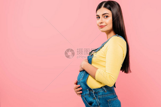 怀孕的漂亮女孩触摸被粉红色隔离的腹部的侧视图图片