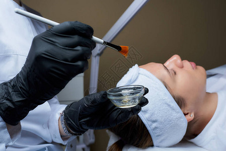一位戴着黑色一次医用手套的女美容师准备用刷子给女孩擦透明的脸皮在美容院进行皮肤清图片