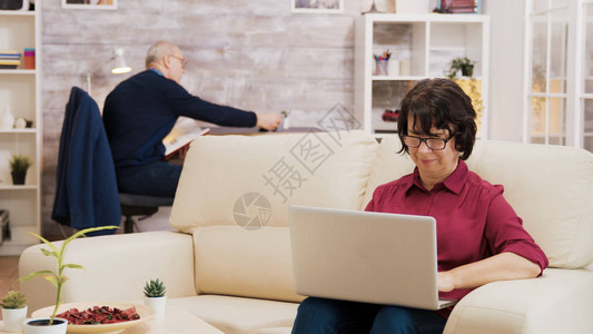身戴眼镜的老年妇女坐在沙发上用笔记本电脑工作图片