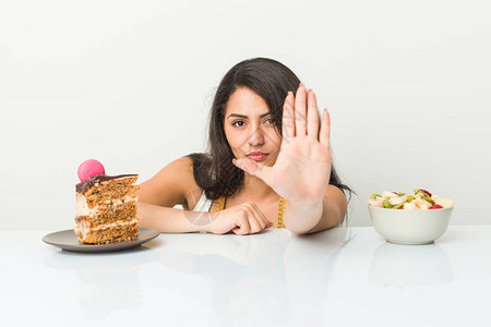 年轻的西班牙女人在蛋糕和水果之间选择手伸展的手举着停止牌阻止你图片