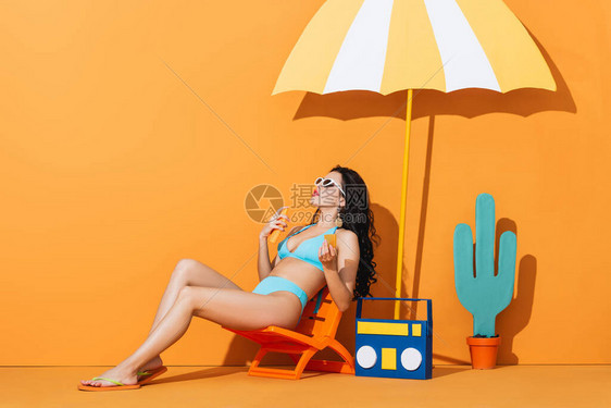 戴太阳眼镜和泳衣的时装女孩坐在纸机仙人掌和雨伞附近的甲板椅上图片