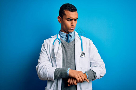 蓝色背景中穿着外套和听诊器的帅气非洲裔美国医生检查手表上的时间图片