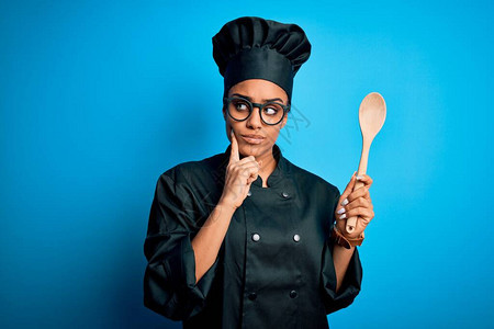 身着烹饪制服的年轻非洲女厨师女孩戴着帽子拿着木勺子工具严肃地面对问题图片