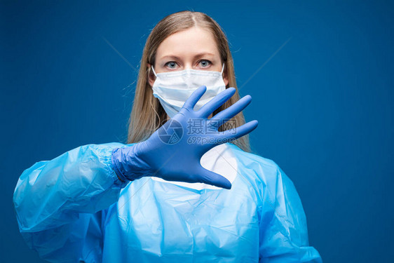 穿着防护服的年轻女子在手套中用手做势保护特写在蓝色工作室背景下隔离的医疗呼吸器和制服图片