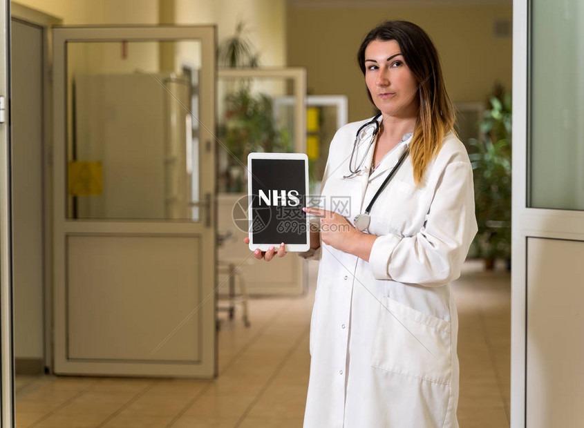 中年白人身穿白袍手持听诊器并指着带有NHS文字的平板电脑的女医生图片