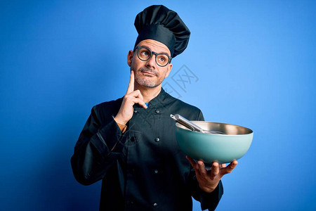 年轻英俊的厨师穿着炊具制服和帽子混合用搅拌器和碗严肃的脸思考问题图片