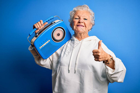拿着老式收音机的资深美女站在孤立的蓝色背景上图片