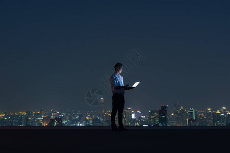 亚洲商人站在大楼的顶部使用笔记本与城市夜景商业成图片