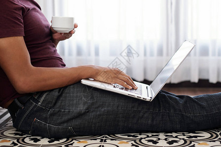 青年男子坐在家中地板上喝早咖啡和答复电子邮件的剪裁图像单图片