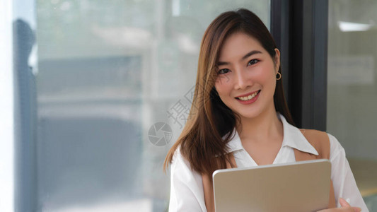 女大学生在站玻璃墙边微笑并持有数字平板电脑的肖像图片