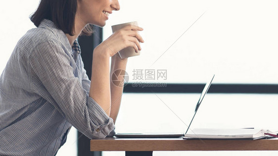 上午咖啡剪裁的幸福学生拿着白咖啡杯观看笔记本电脑录像图片