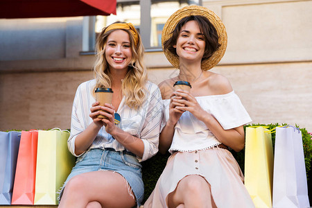 一个快乐高兴的年轻女孩朋友坐在户外喝咖啡在购物袋附近互图片