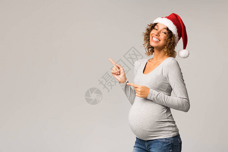 身戴圣塔帽的令人兴奋的Afro孕妇指着灰色背图片