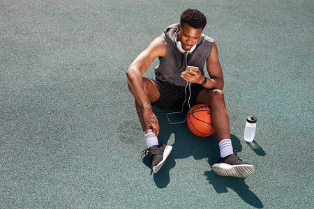 坐在篮球场并使用智能手机复制空间的英俊非洲裔美国人男子的背景请查图片
